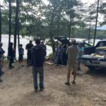 a group of people standing in a circle in front of the water with vehicles carrying equipment
