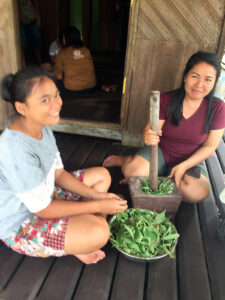Two smiling women process vegetation with a mortar and pestle
