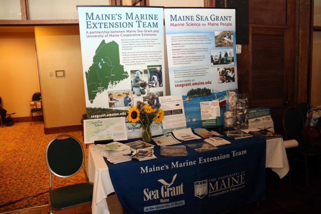 A display table holds 2 Maine Sea Grant banners, a bouquet of flowers, and information pamphlets and handouts.