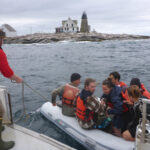 People wearing lifejackets siting in zodiak as it gets attached to a larger vessel.