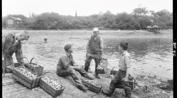 A man and a boy, each holding a basket full of shellfish, talk to a man sitting on the bank of New Harbor. Another man stands to the left and is looking down at multiple baskets of shellfish. Photo credit Maine State Archives.