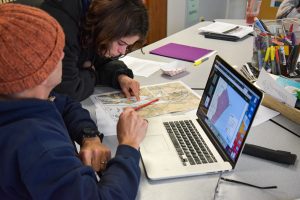 two people at a computer looking at paper maps on a cluttered desk