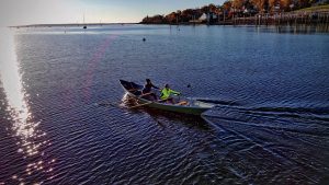 Two people rowing in a boat on the water with docks and houses on shore in the background
