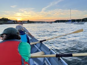 three people in a boat with oars, with the sun on the horizon in the background