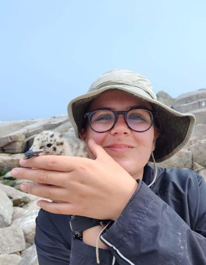 a person in a hat on the rocky coast holding a juvenile seabird.