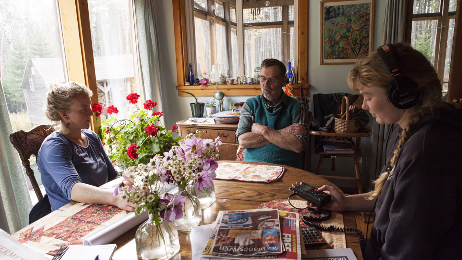 Three people sit around a dining room table with three vases of wildflowers in the center. On the right, a student with headphones uses a hand-held recording device to document the stories of a man in a green vest. Another student sits at the table with them, listening.
