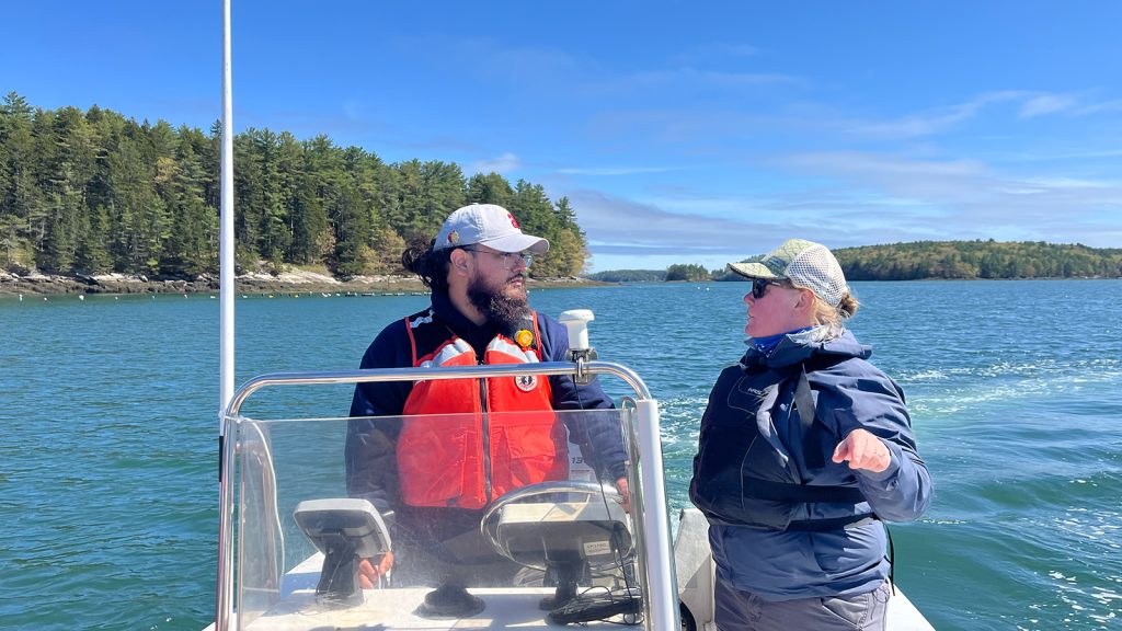 A man driving a boat on a beautiful sunny day listens to his coach as she teaches.