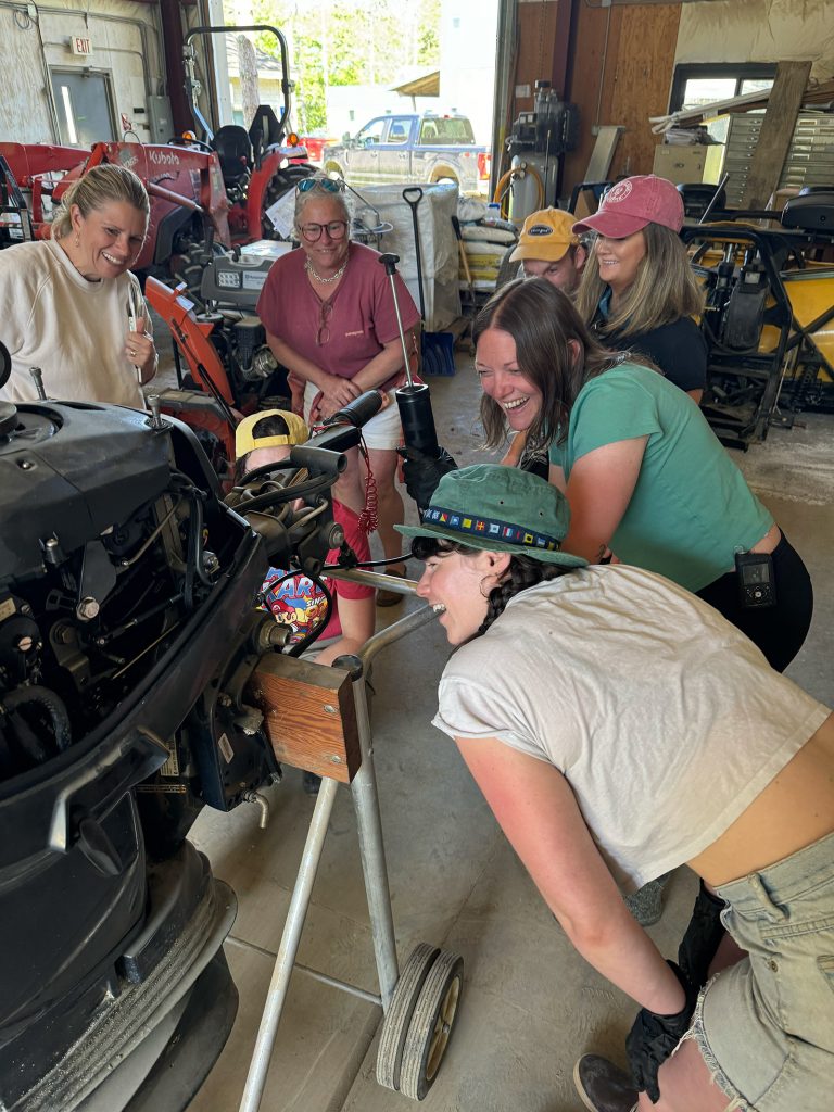 Several people giggle as they gather around an outboard engine together