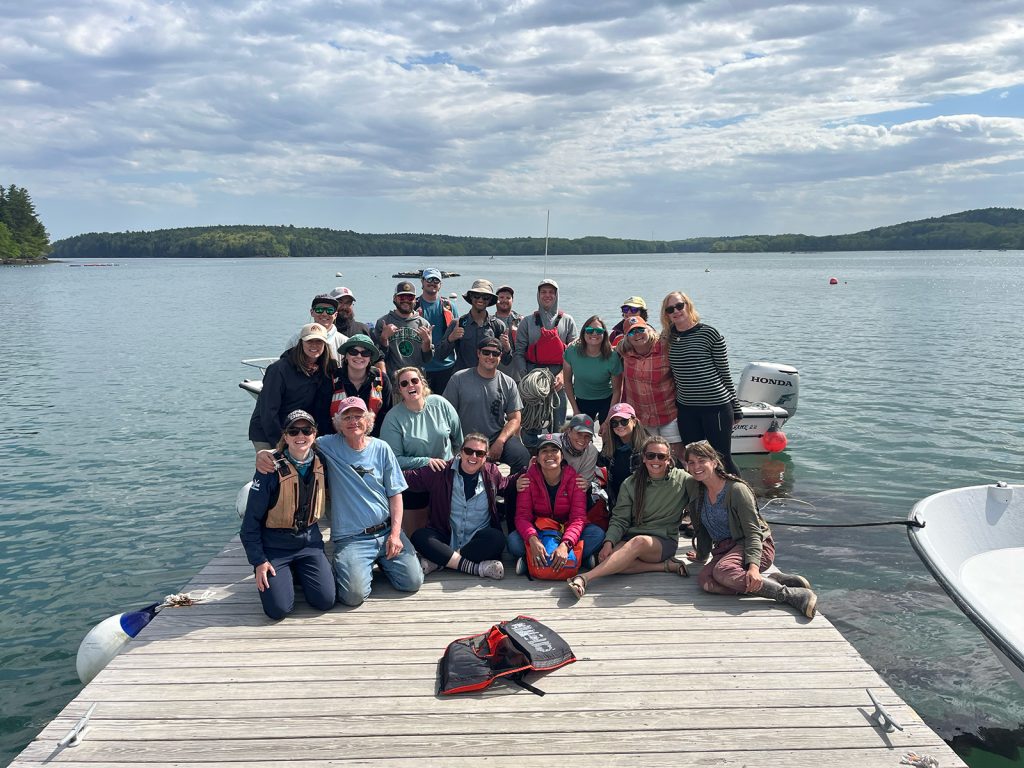 A group of smiling people pose on a dock with water and fairweather clouds behind them