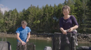 A woman and a man standing by the water holding black plastic shellfish bags