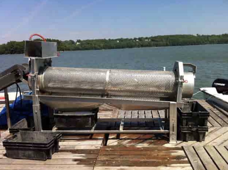 a long perforated metal cylinder is mounted horizontally on a metal frame. A motor is attached to one end and a container to catch oysters is on the pier on the other end